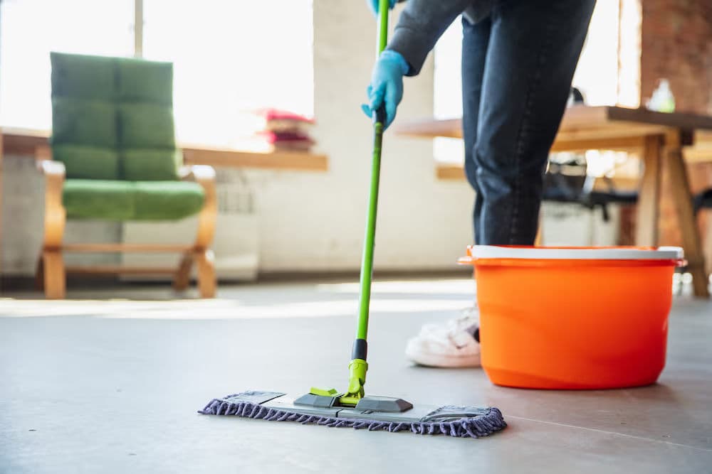 Person cleaning floor with microfiber mop and orange bucket in bright, modern room.